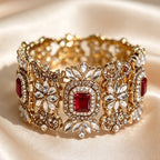 An ornate gold-toned cuff bracelet with red gemstone accents and clear crystal floral detailing, displayed on draped cream satin fabric.